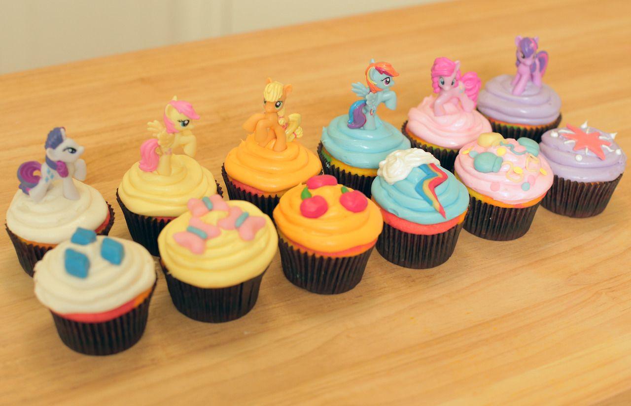 A row of decorated cupcakes topped with colorful frosting and pony figurines, featuring various themes and vibrant designs.
