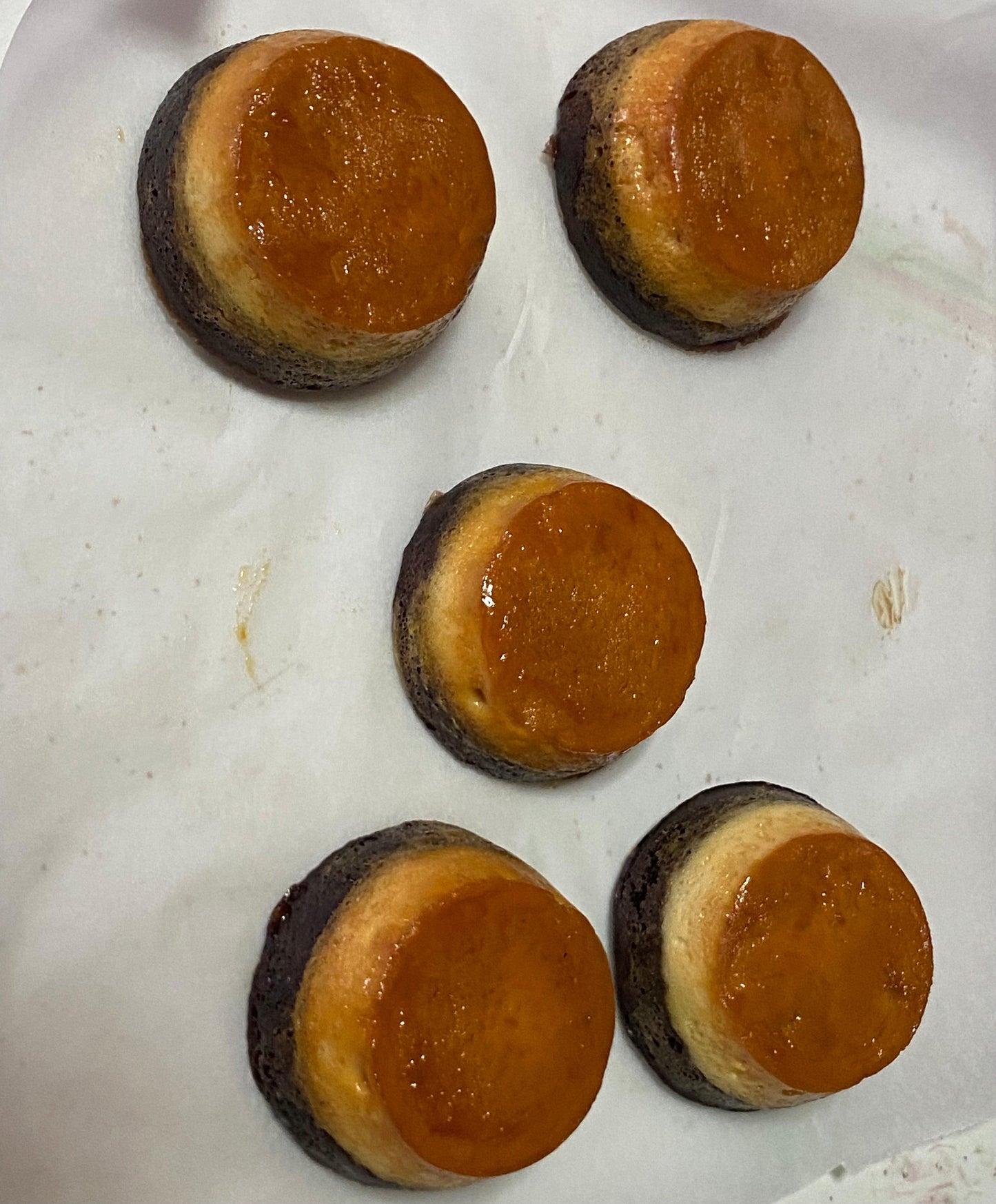 Four round pastries with a glossy orange glaze on top, resting on white parchment paper. The bases are dark brown with a light edge.