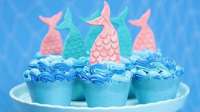 A tiered cake stand features blue cupcakes topped with colorful mermaid tails,