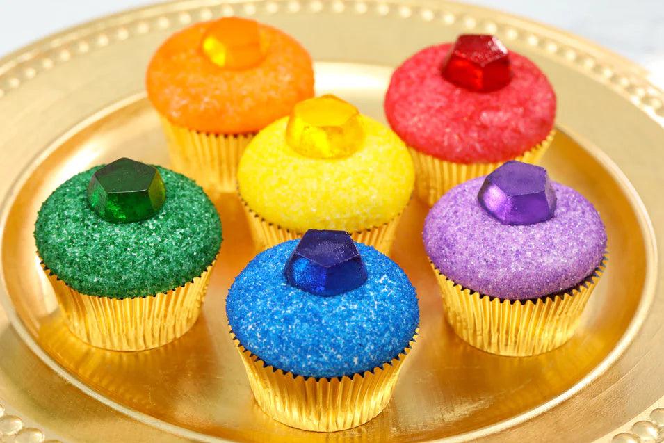 A golden platter displays six colorful cupcakes, each topped with sparkling sugar and a gem-like candy in various vibrant colors.