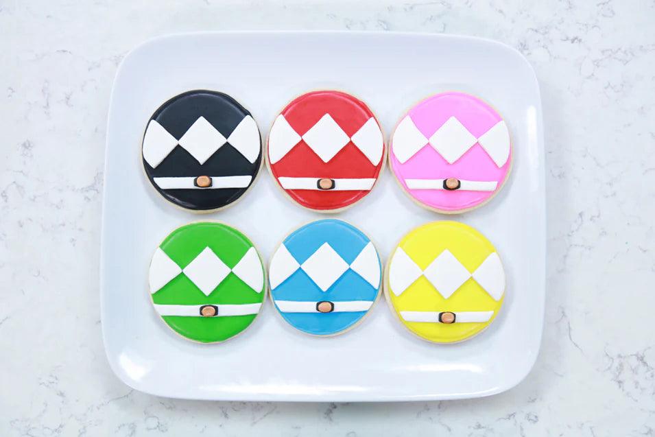A plate of six cookies decorated as colorful Power Ranger helmets: black, red, pink, green, blue, and yellow