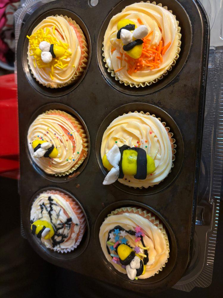 A tray of six colorful cupcakes topped with cream, playful bee decorations, and vibrant sprinkles.