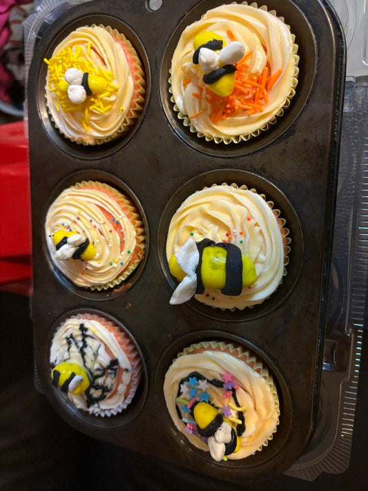 A tray of six colorful cupcakes topped with cream, playful bee decorations, and vibrant sprinkles.